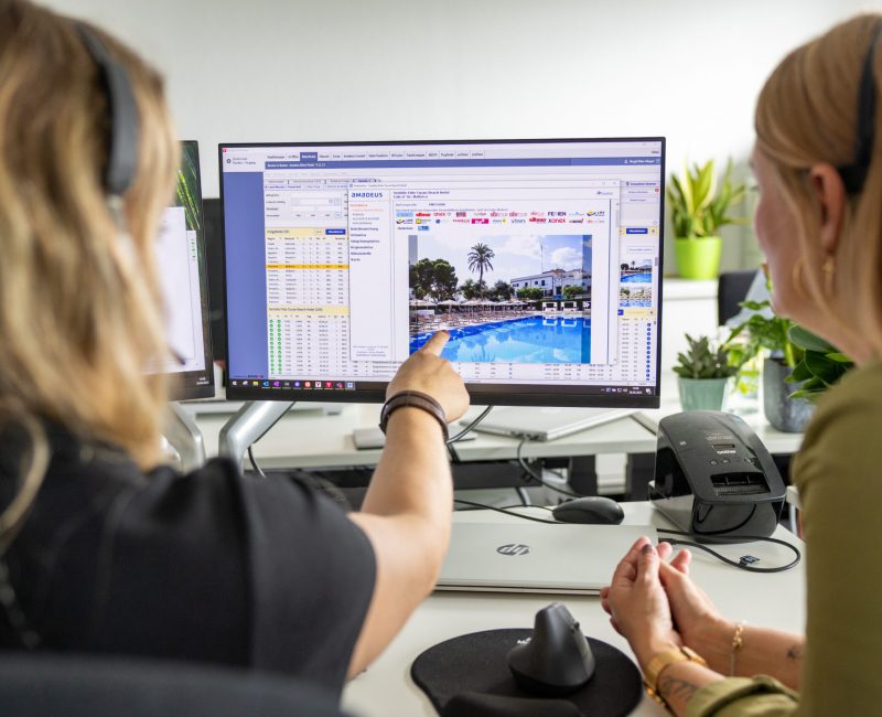 Zwei Personen sitzen vor einem PC und eine zeigt auf dem Bildschirm, wo das CRS System AMADEUS angezeigt ist